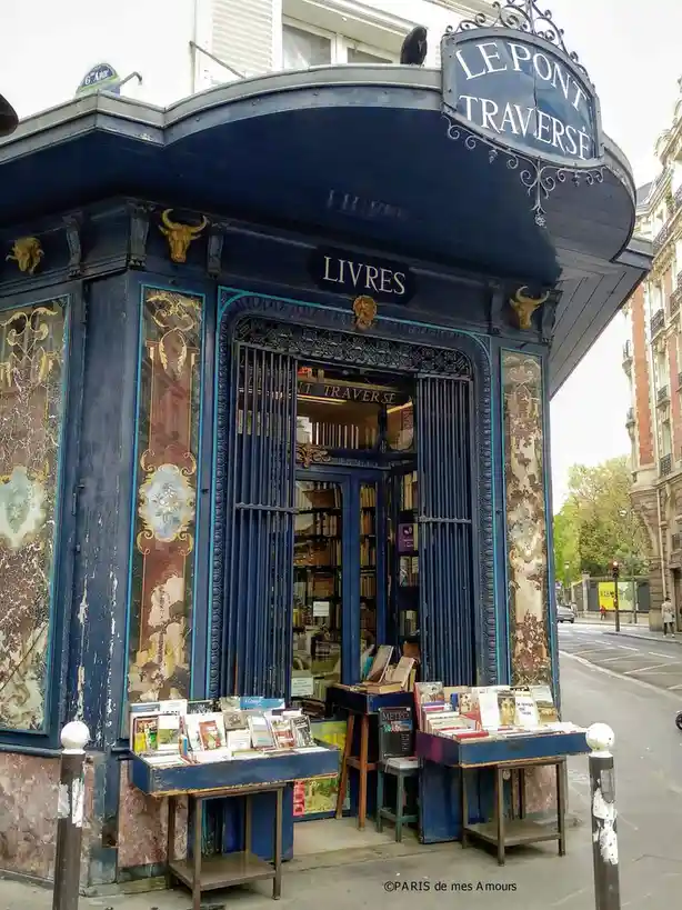An ornate, blue-painted Parisian bookstore named 'LE PONT TRAVERSÉ' with 'LIVRES' above the door, displaying books on tables…