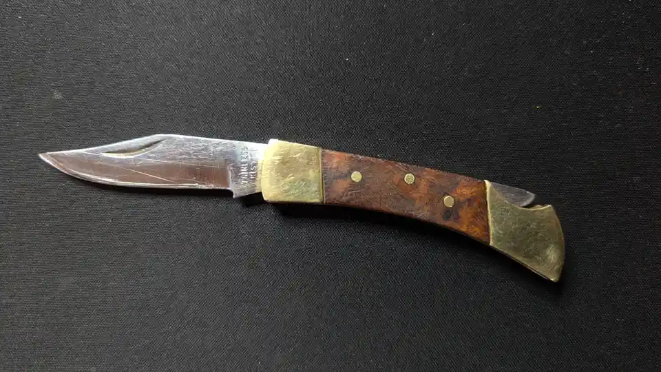 A close-up photo of a small, worn pocket knife with a stainless steel blade and a brown wooden handle secured by brass…