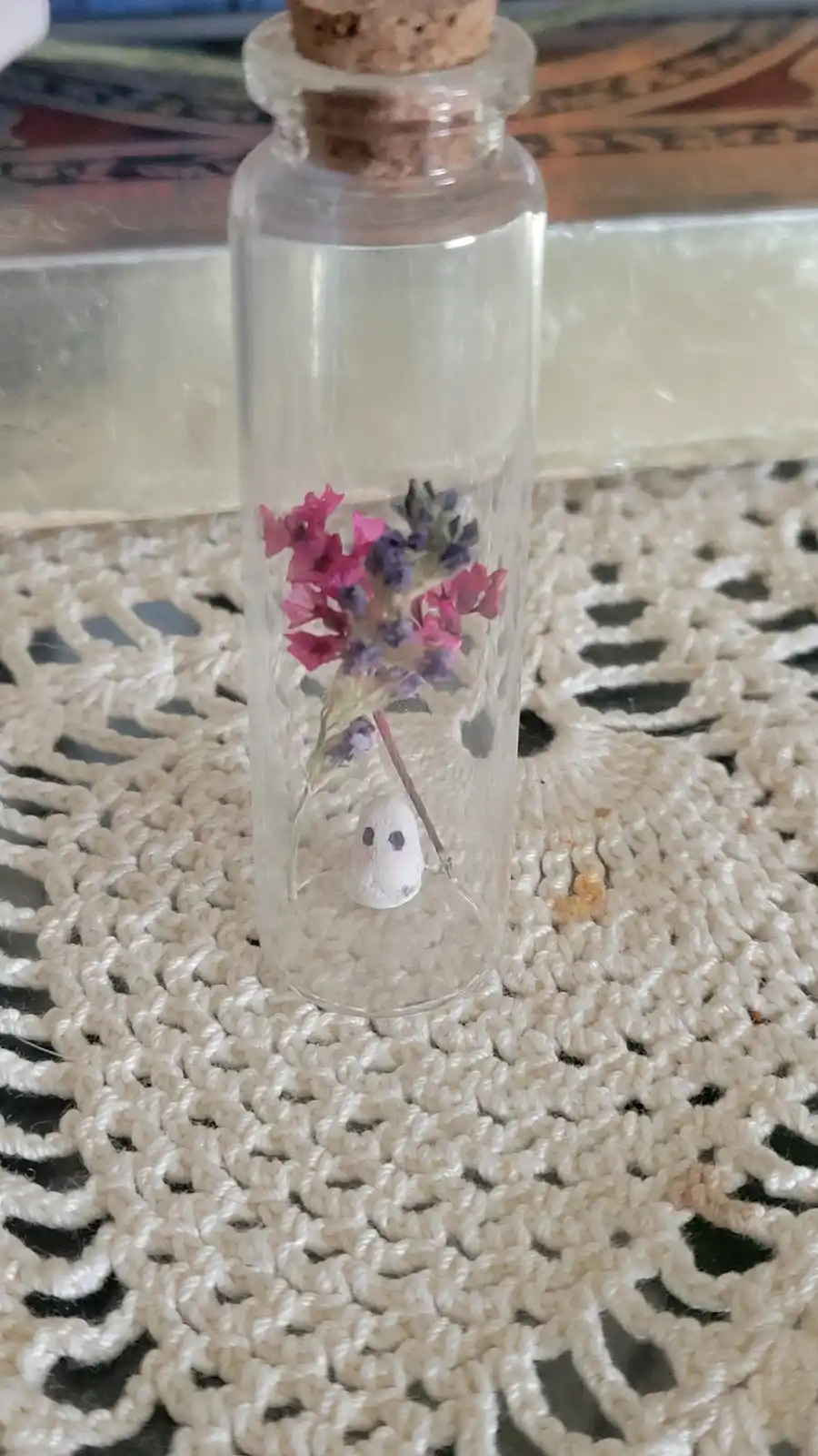 A small glass bottle with a cork stopper contains dried pink and purple flowers and a tiny white ghost figurine with black…
