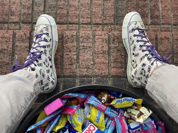 A person wearing white sneakers covered in Joker face and 'HA! HA! HA!' text sits on a brick sidewalk with a black bowl full…