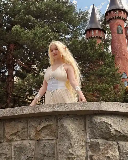 A person with long blonde hair, pointed ears, and wearing a tiara, a white corseted dress, and silver arm cuffs stands on a…