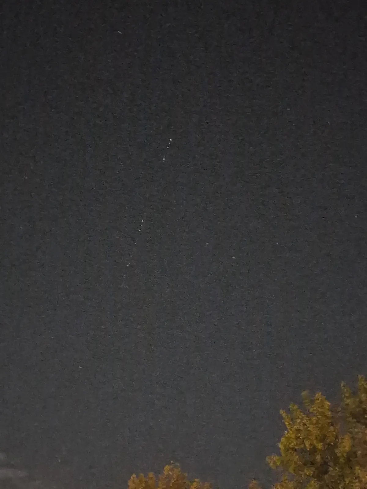 A dark night sky with faint stars and a vertical line of bright dots, possibly a meteor trail or camera artifact, with the…