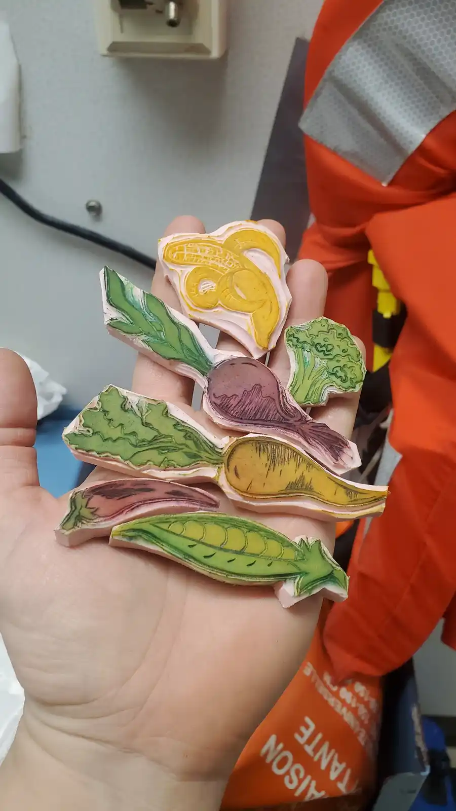 A hand holds several hand-carved wooden stamps shaped like vegetables including corn, broccoli, beet, carrot, eggplant, and…
