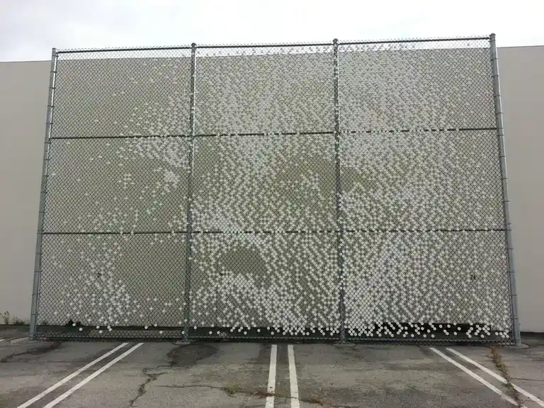 a large chain-link fence in a parking lot is covered with numerous white dots arranged to form a recognizable image of a…