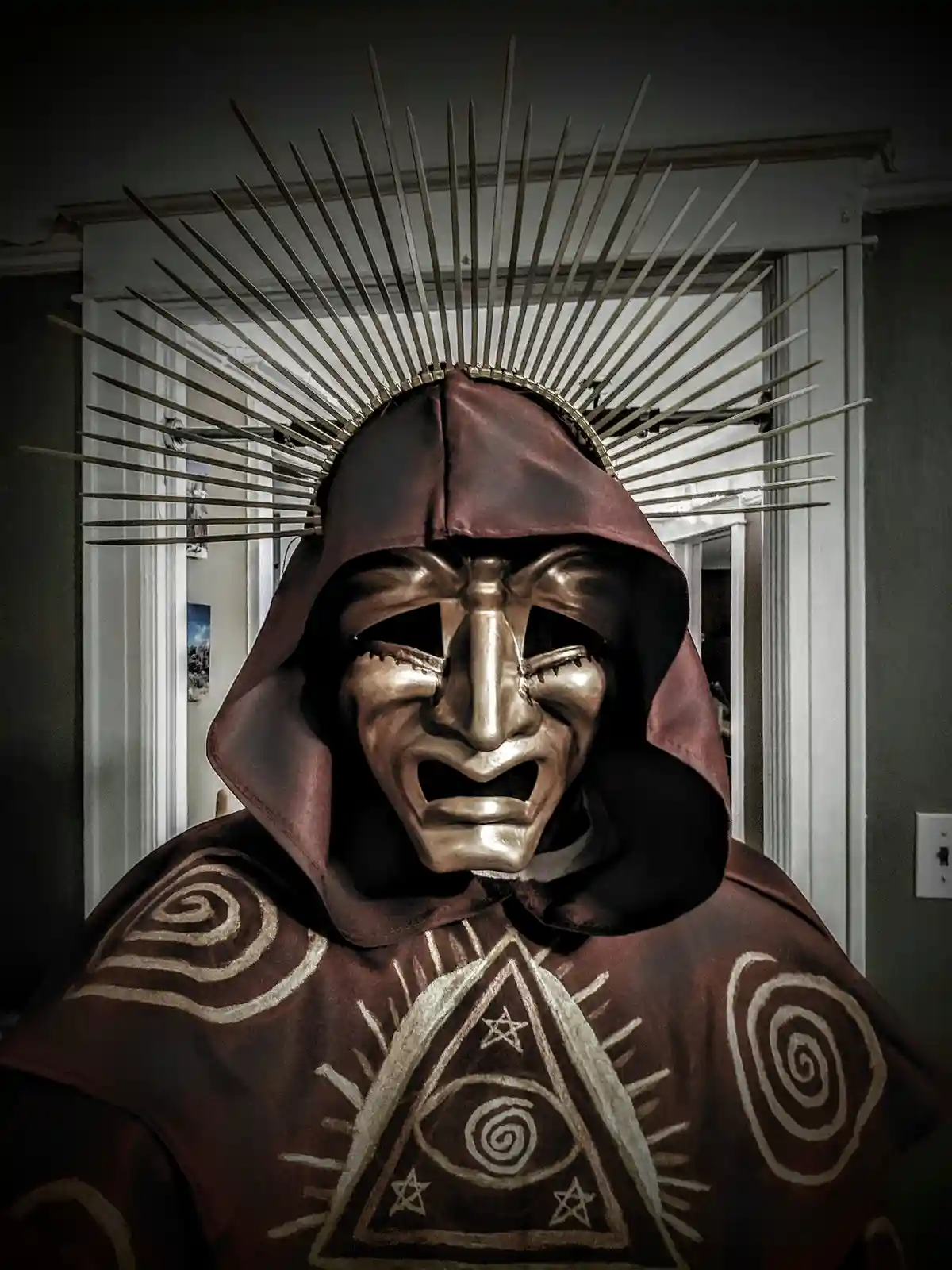 A person wearing a dark red hooded robe with occult symbols, including an all-seeing eye within a triangle and spirals, and…