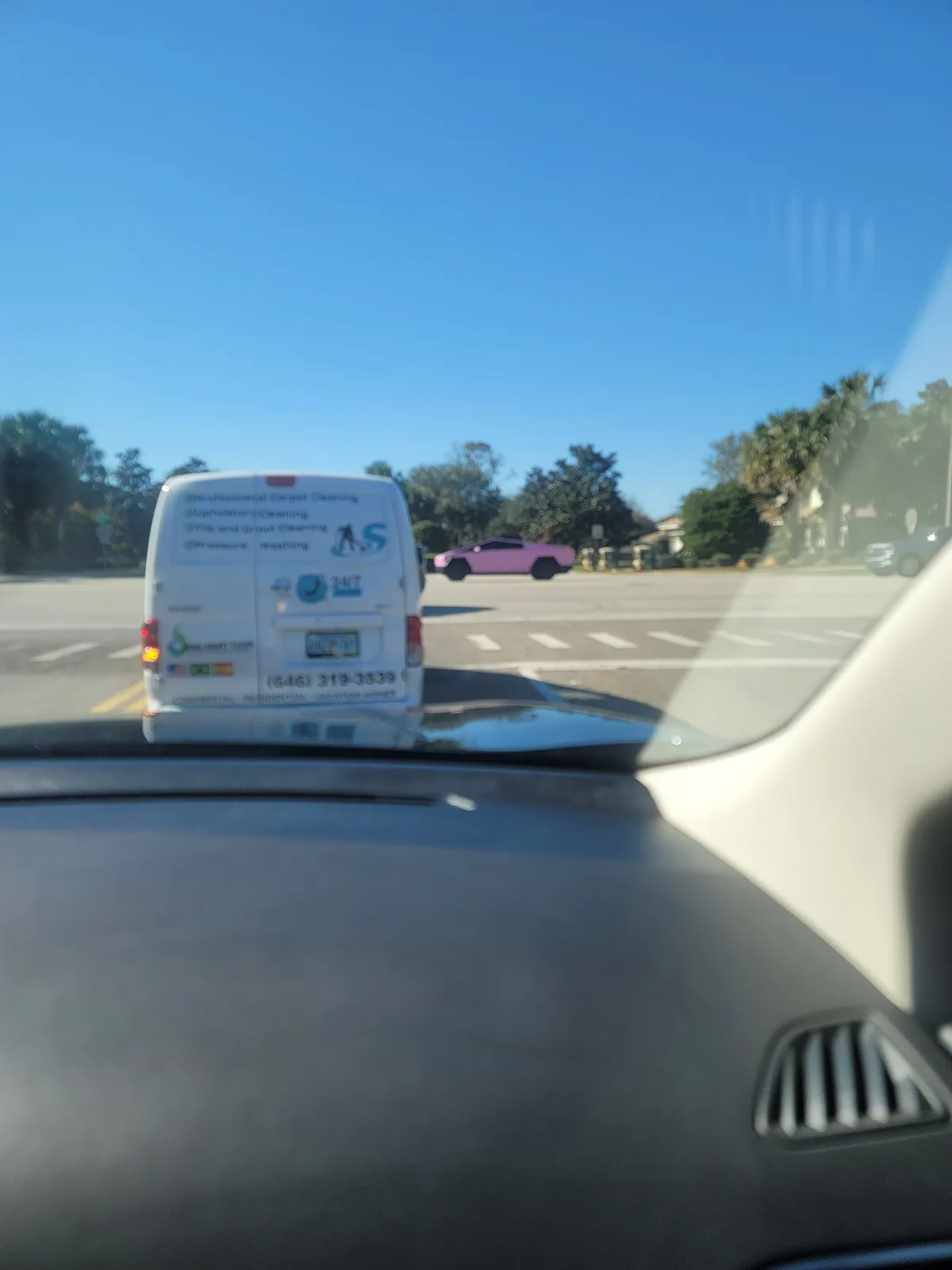 A photo taken from inside a car shows a white van with cleaning service ads in front, while a bright pink car is visible in…