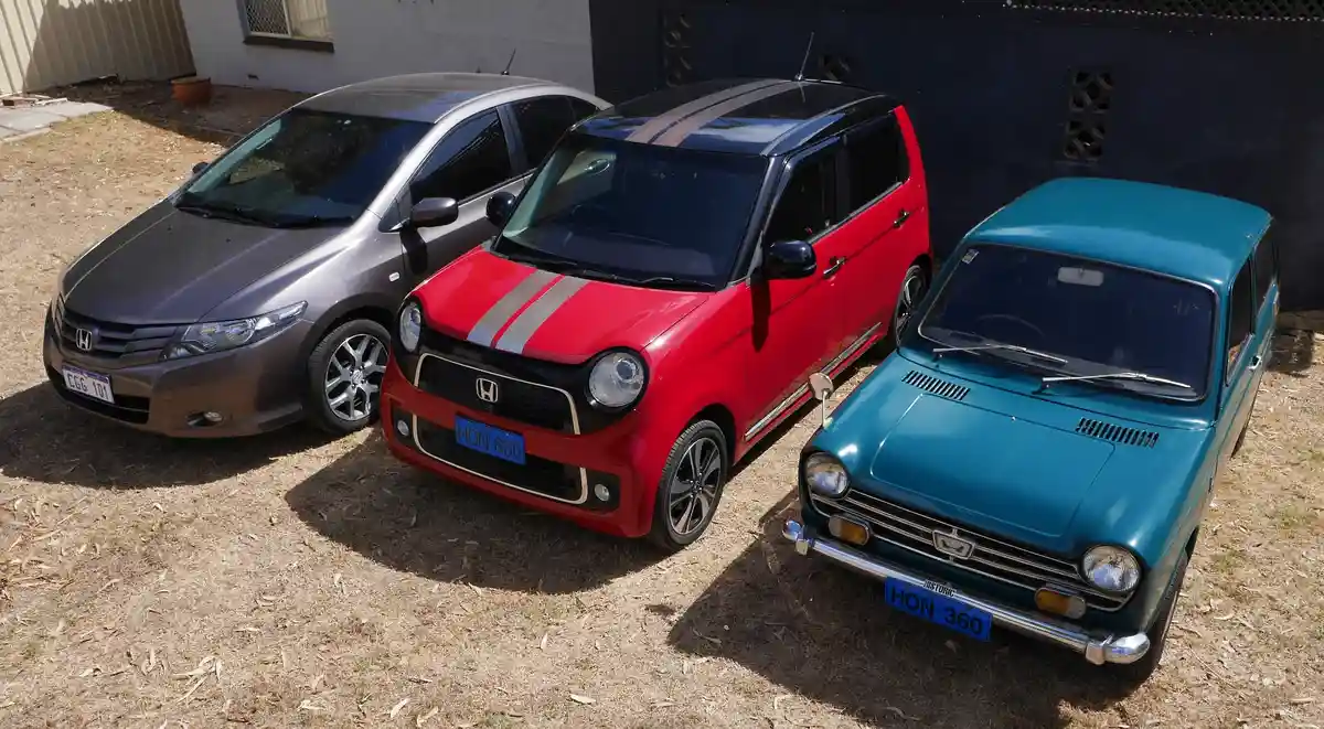 Three Honda cars of different eras are parked side-by-side on dry grass: a modern grey sedan, a red compact hatchback with…