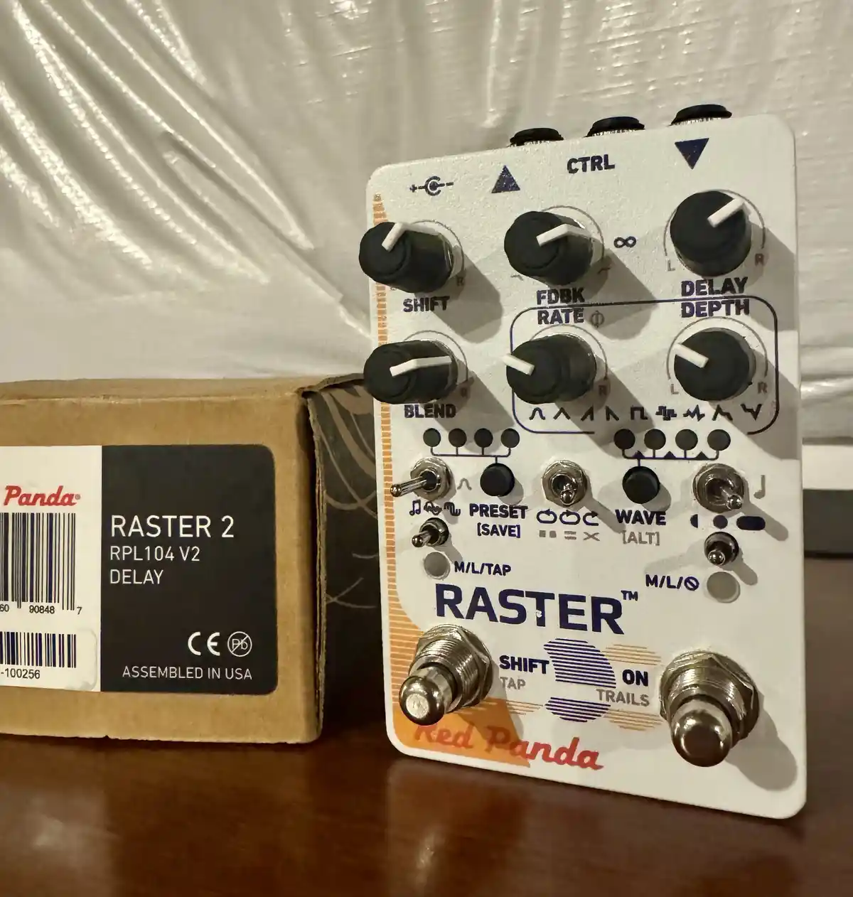 A white Red Panda Raster 2 delay pedal with black knobs and silver footswitches sits next to its cardboard box labeled…