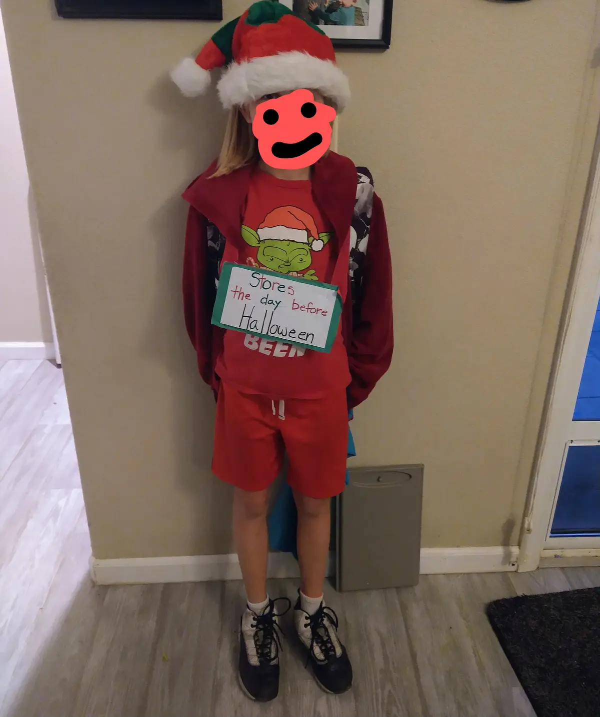 A child wearing a red Santa hat and red holiday-themed clothing stands indoors holding a handwritten sign that reads 'Stores…