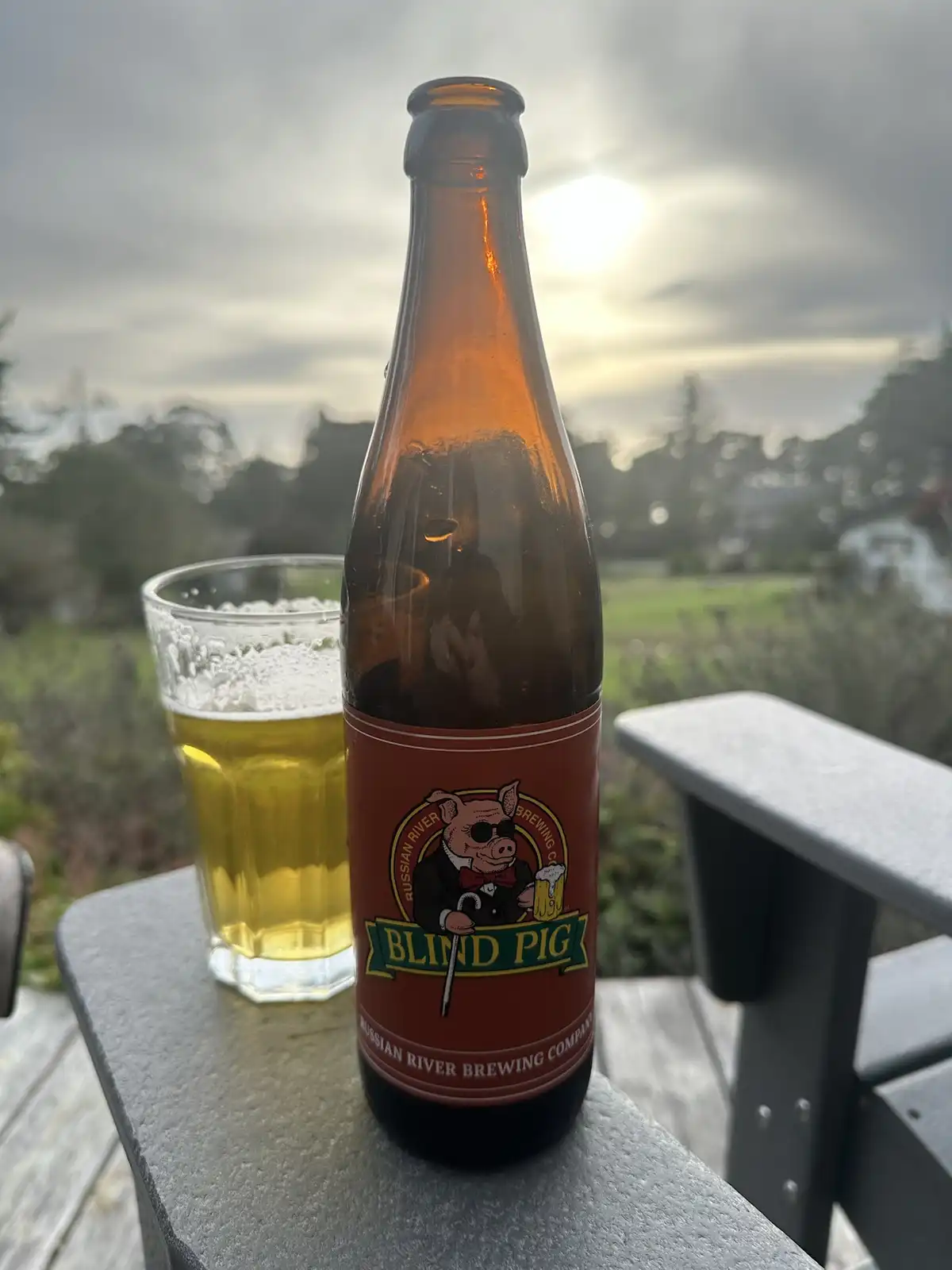 A brown beer bottle labeled 'Blind Pig' from Russian River Brewing Co. sits on a gray outdoor table next to a glass of beer…