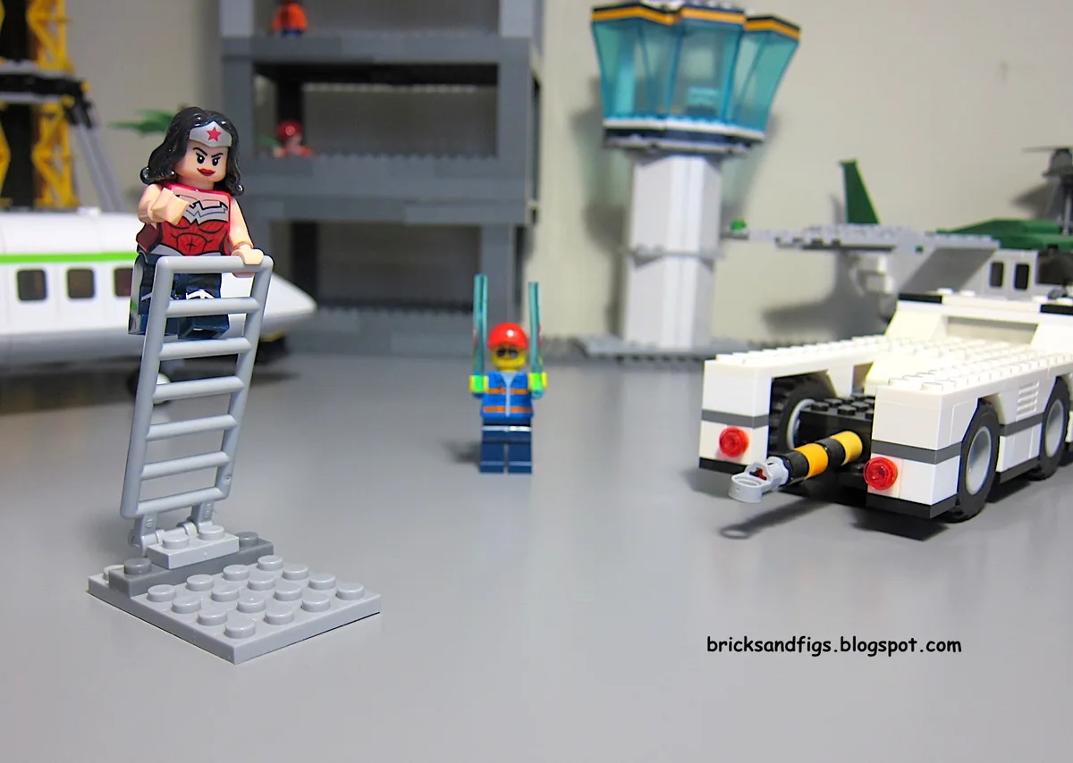 A lego wonder woman minifigure stands atop a grey ladder on a grey baseplate, facing a lego airport worker holding two green…