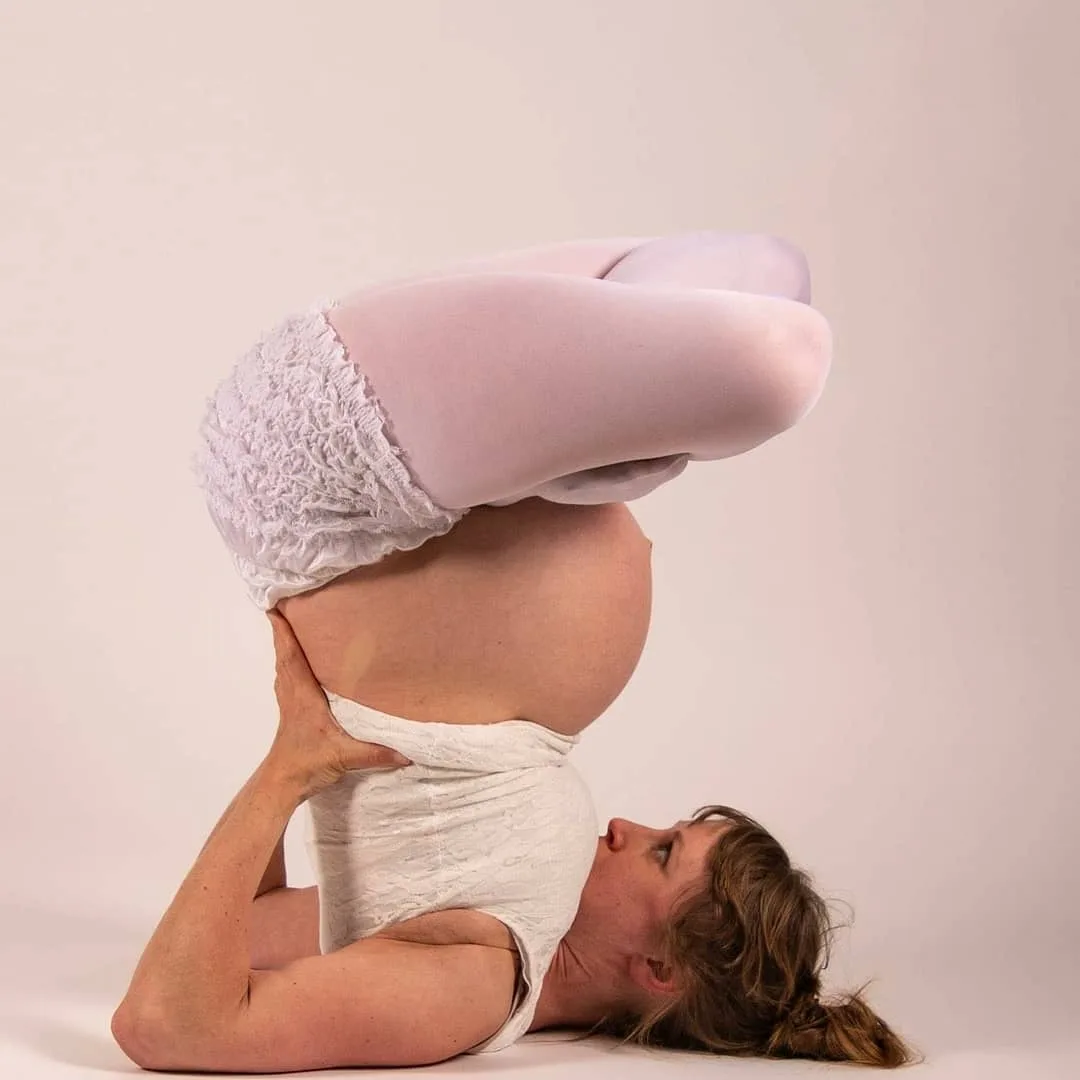 A pregnant woman is performing a headstand yoga pose against a plain background, her legs bent at the knees and feet…