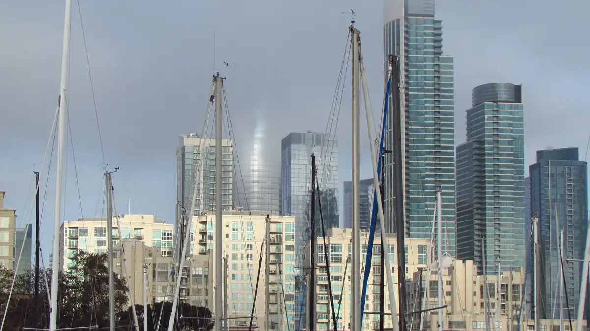 A city skyline with modern glass skyscrapers is viewed through the masts of sailboats in a harbor. One central skyscraper is…