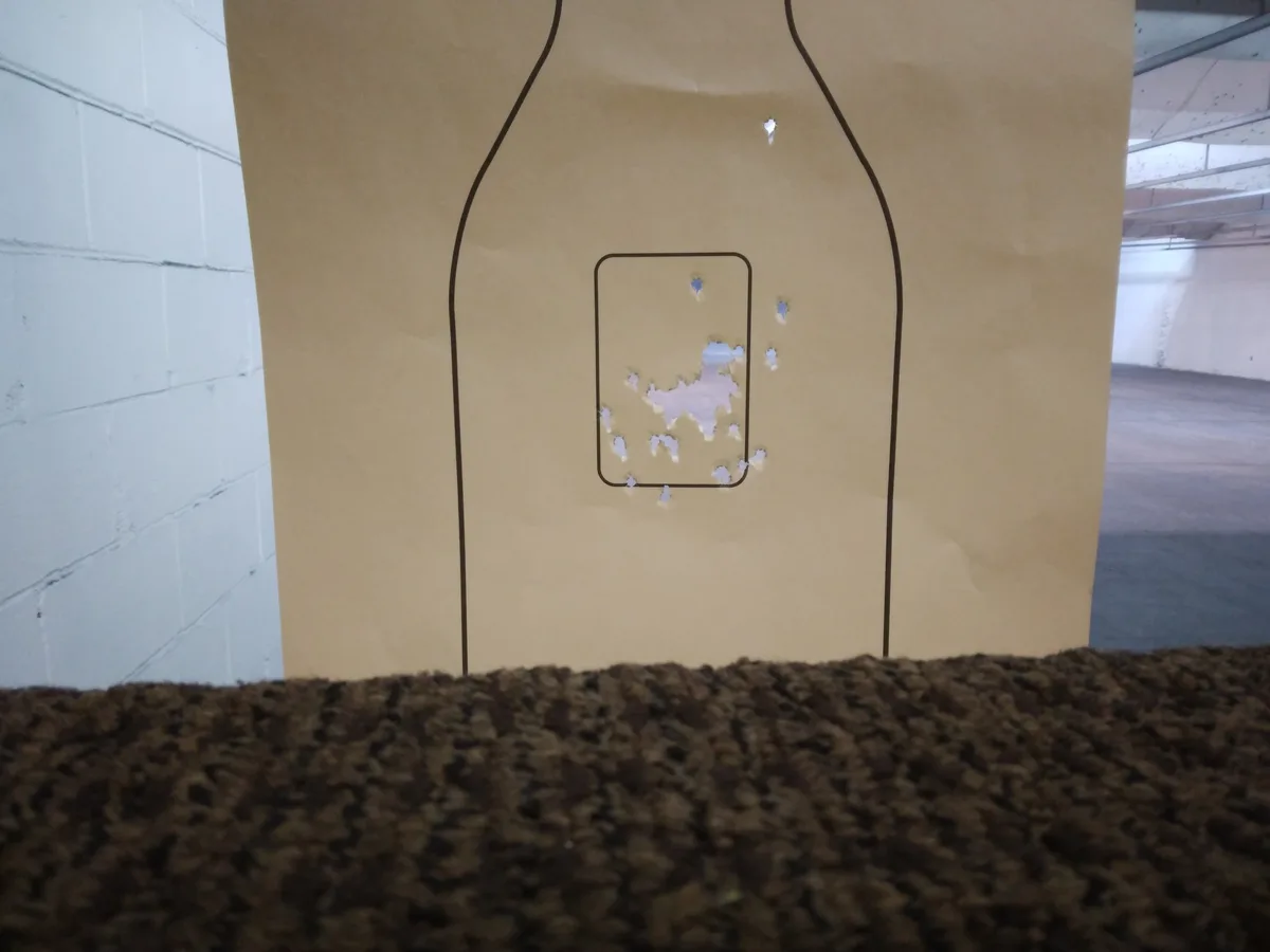 A tan paper target shaped like a bottle with a rectangular outline drawn on its body, showing numerous bullet holes…