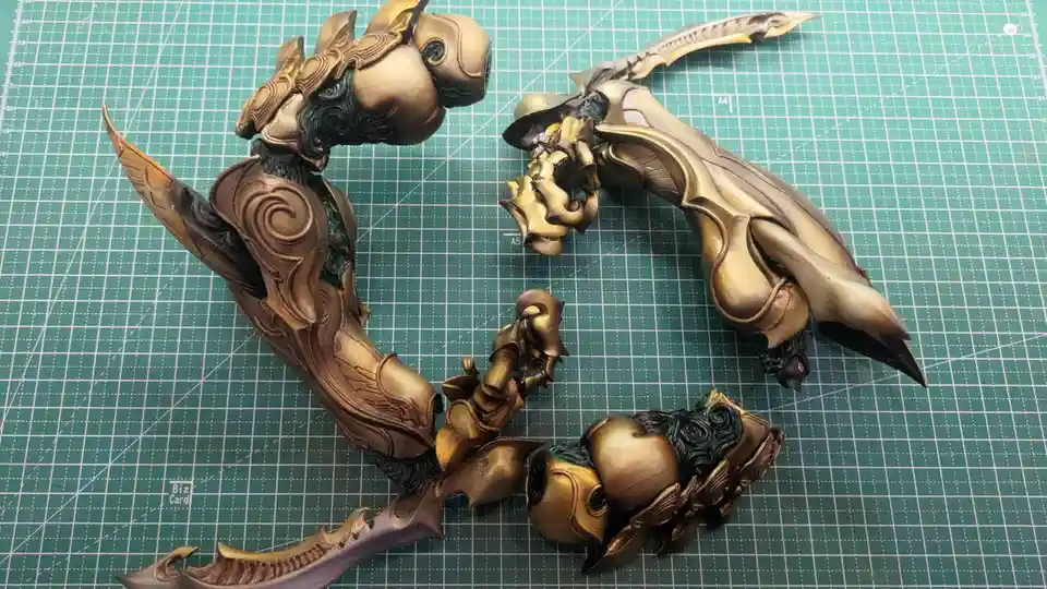 Two intricately painted, golden-brown fantasy weapon or armor pieces lie scattered on a green cutting mat with grid lines…