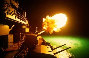 A naval gun on a ship fires a bright orange shell into the dark water, illuminating the scene with a greenish glow. The…