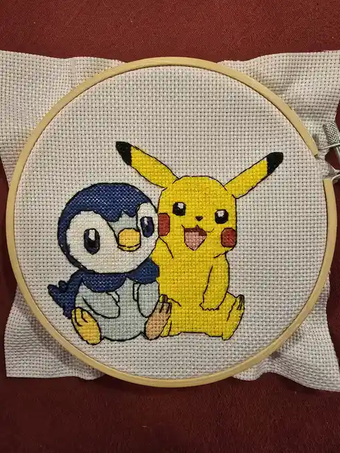 A cross-stitch embroidery in a wooden hoop shows Pikachu and Piplup sitting together on white fabric, with Pikachu on the…