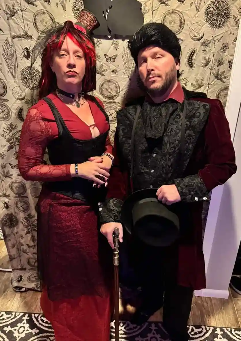 Two people dressed in elaborate gothic or Victorian-style costumes pose together. The woman wears a red lace dress with…