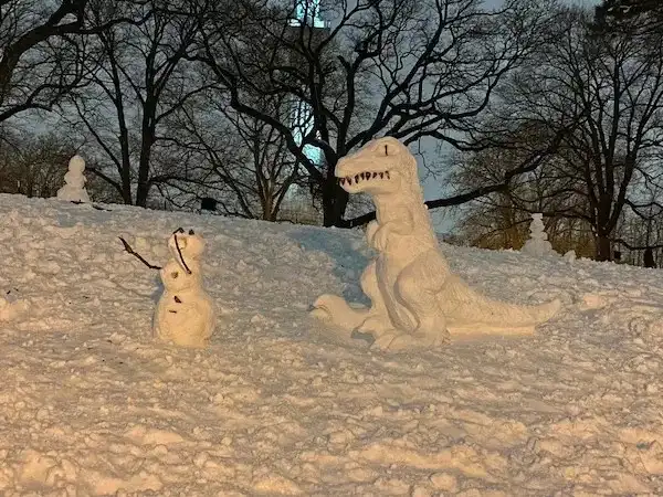 A snowman stands facing a large snow sculpture of a T-Rex, which is positioned as if about to attack. The snowman has a…