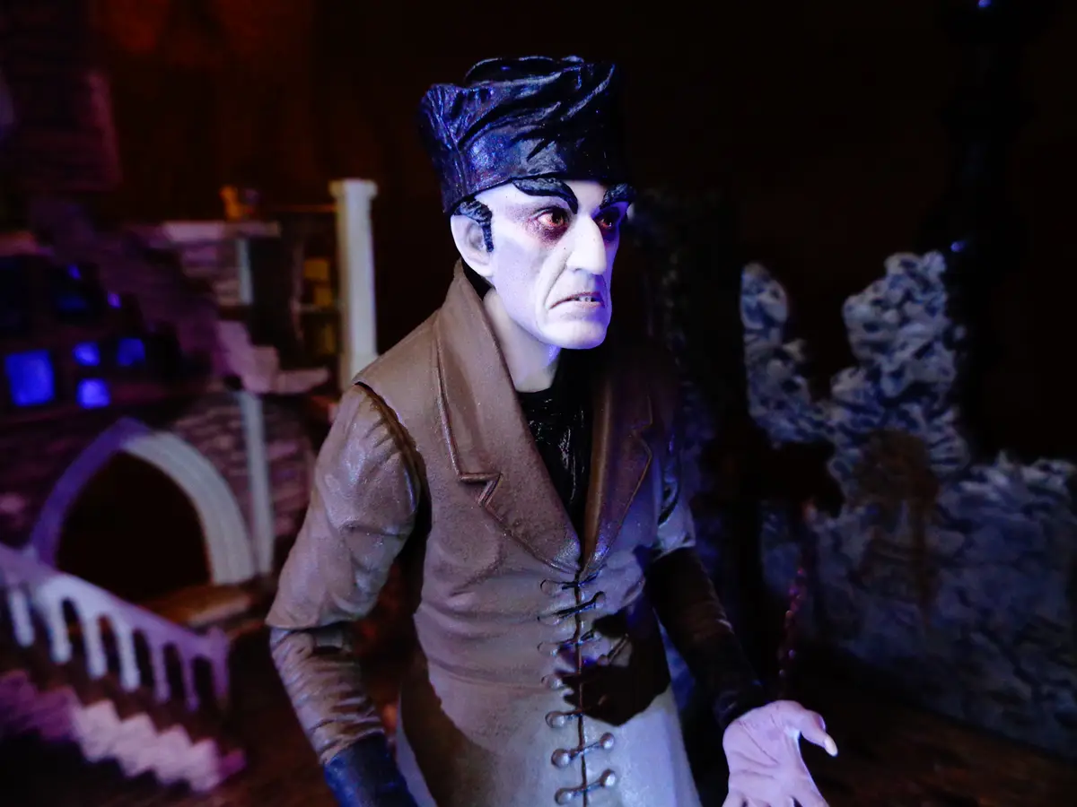 A close-up of a figurine resembling Count Dracula, with pale skin, dark eyes, and a black hat, wearing a brown vest over a…