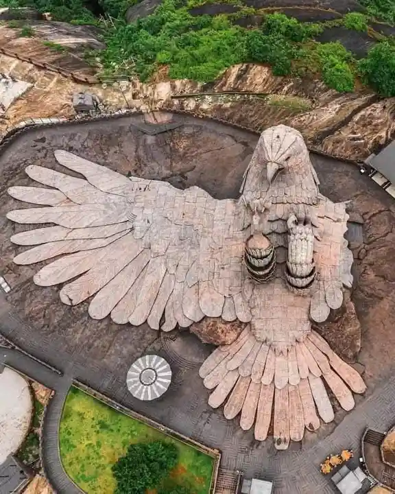 An aerial view shows a massive, stone-tiled sculpture of a bird with wings spread wide, perched on a rocky landscape with…