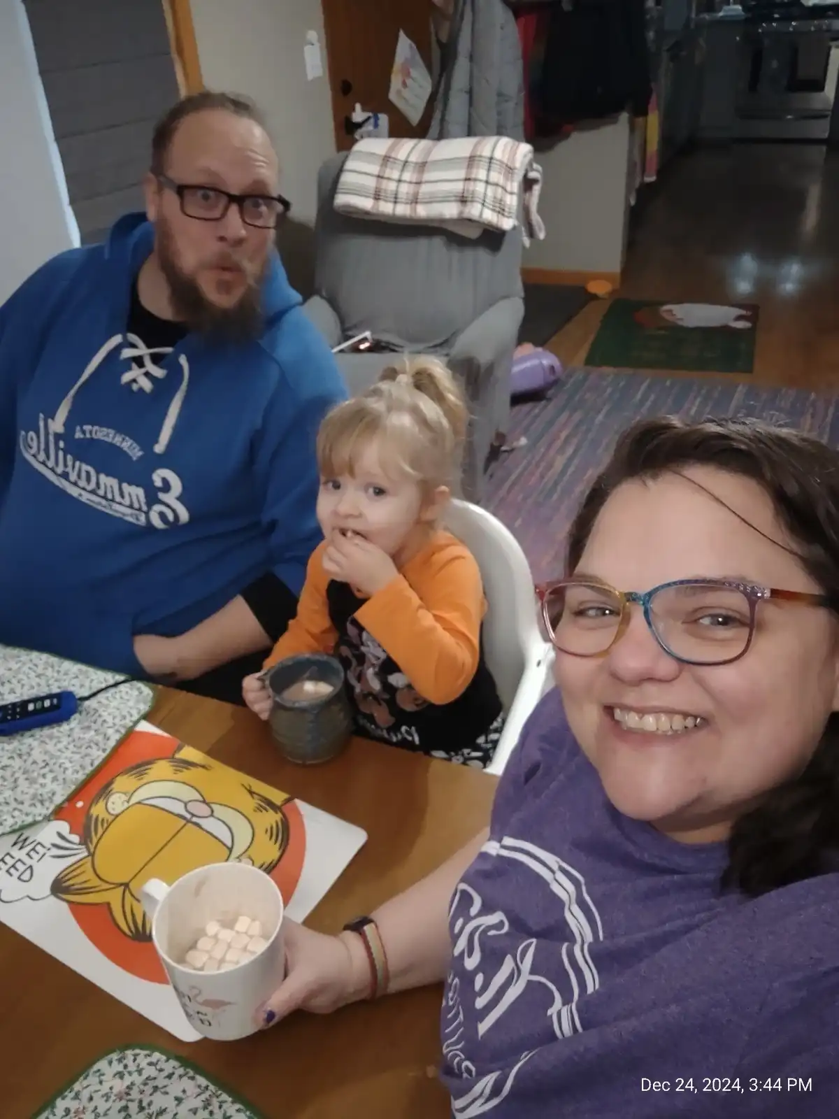 A selfie-style photo shows three people at a wooden dining table in a home. In the foreground, a woman with glasses smiles…