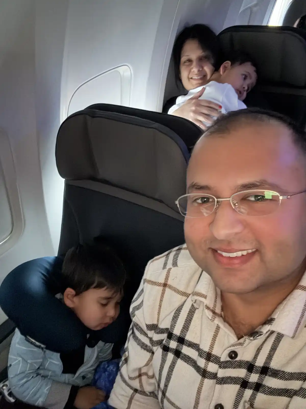 A man in glasses takes a selfie on an airplane, smiling at the camera while two young children sit beside him—one wearing a…