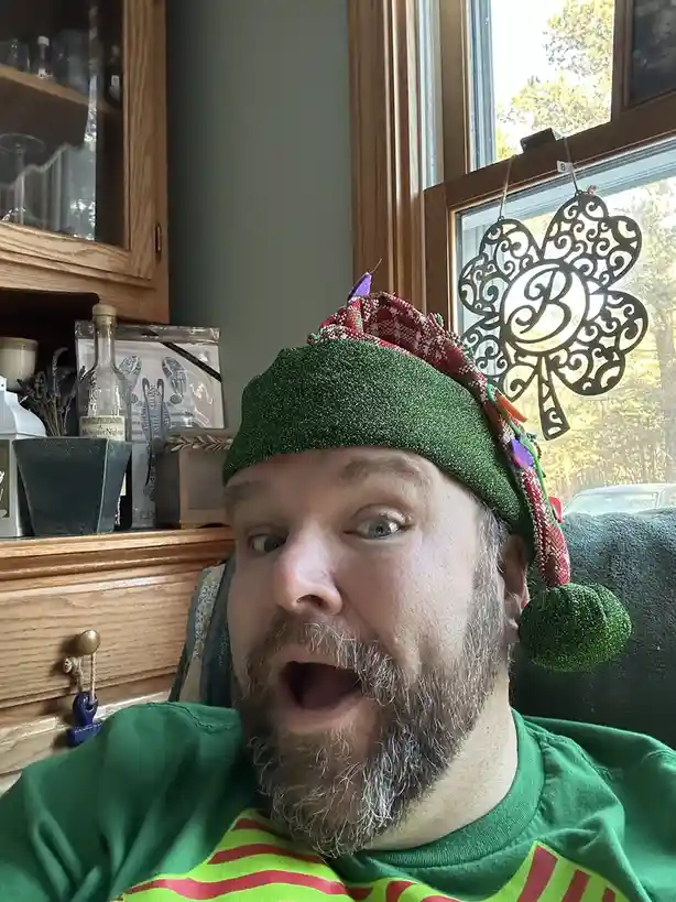 A man with a beard and wide-eyed, open-mouthed expression wears a sparkly green elf hat with red trim and a green pom-pom…