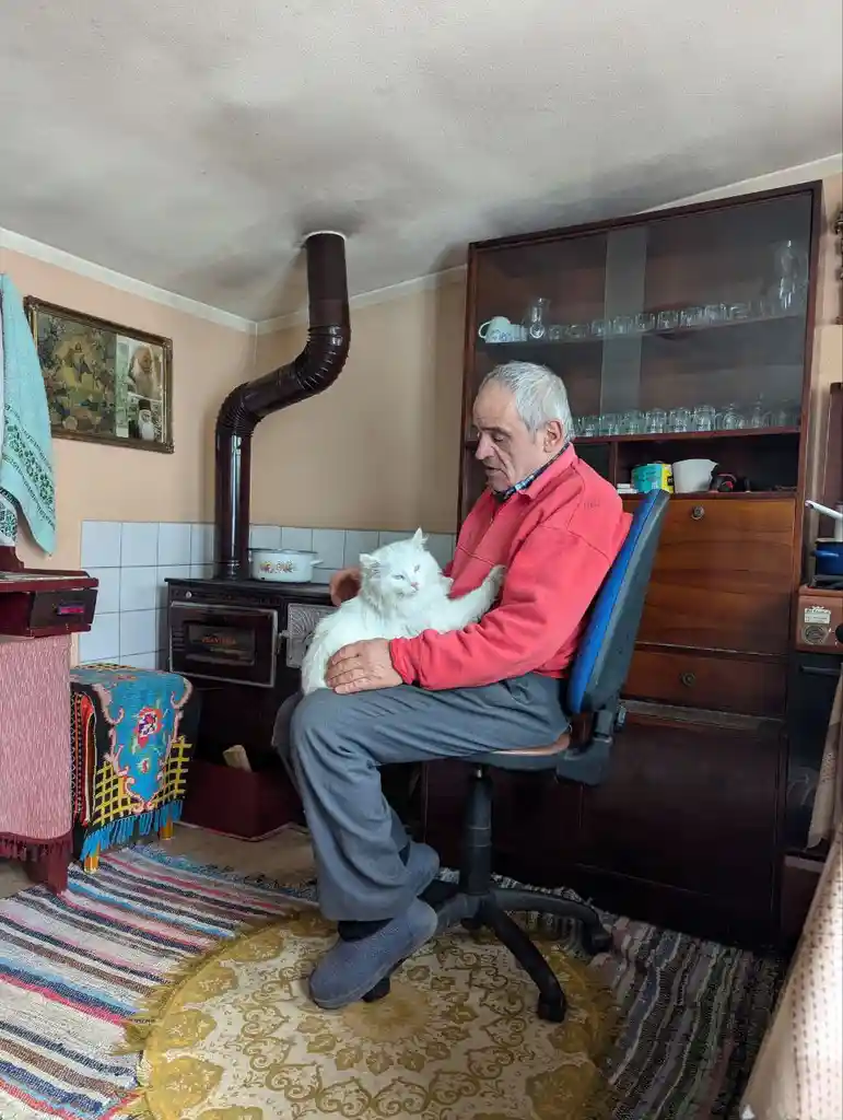 An older man with gray hair sits in a blue office chair in a cozy, slightly cluttered room, holding a fluffy white cat. He…