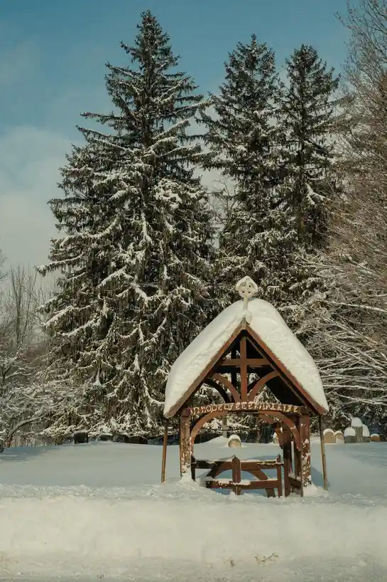 A snow-covered wooden chapel with a cross on top stands in a snowy cemetery, surrounded by evergreen trees. The chapel's…
