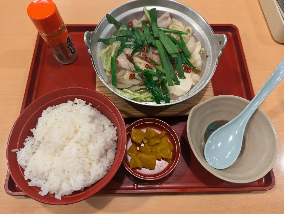 a japanese meal on a red tray includes a metal bowl of soup with noodles, meat, cabbage, green onions, and chili, a bowl of…