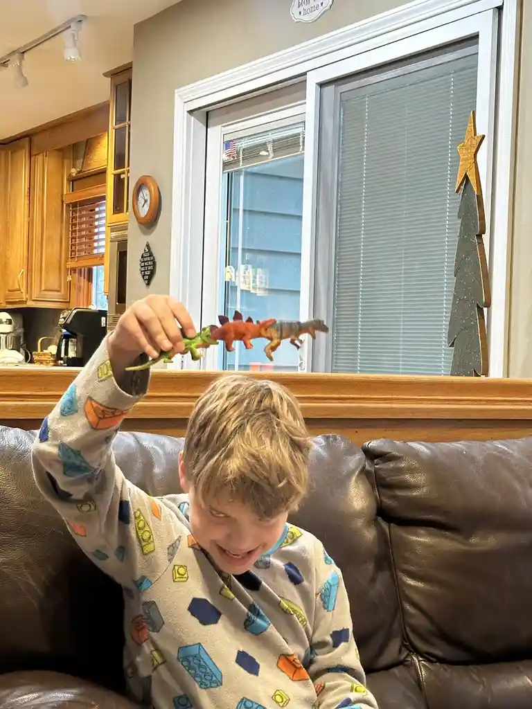 A young boy in lego-patterned pajamas sits on a brown leather couch, smiling as he holds a green plastic dinosaur toy up to…