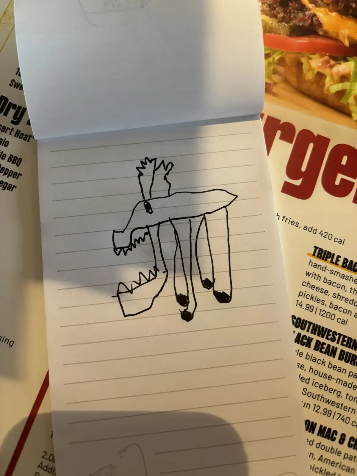 A child's simple black ink drawing on lined notebook paper depicts a creature with a burger patty head, two sharp teeth…