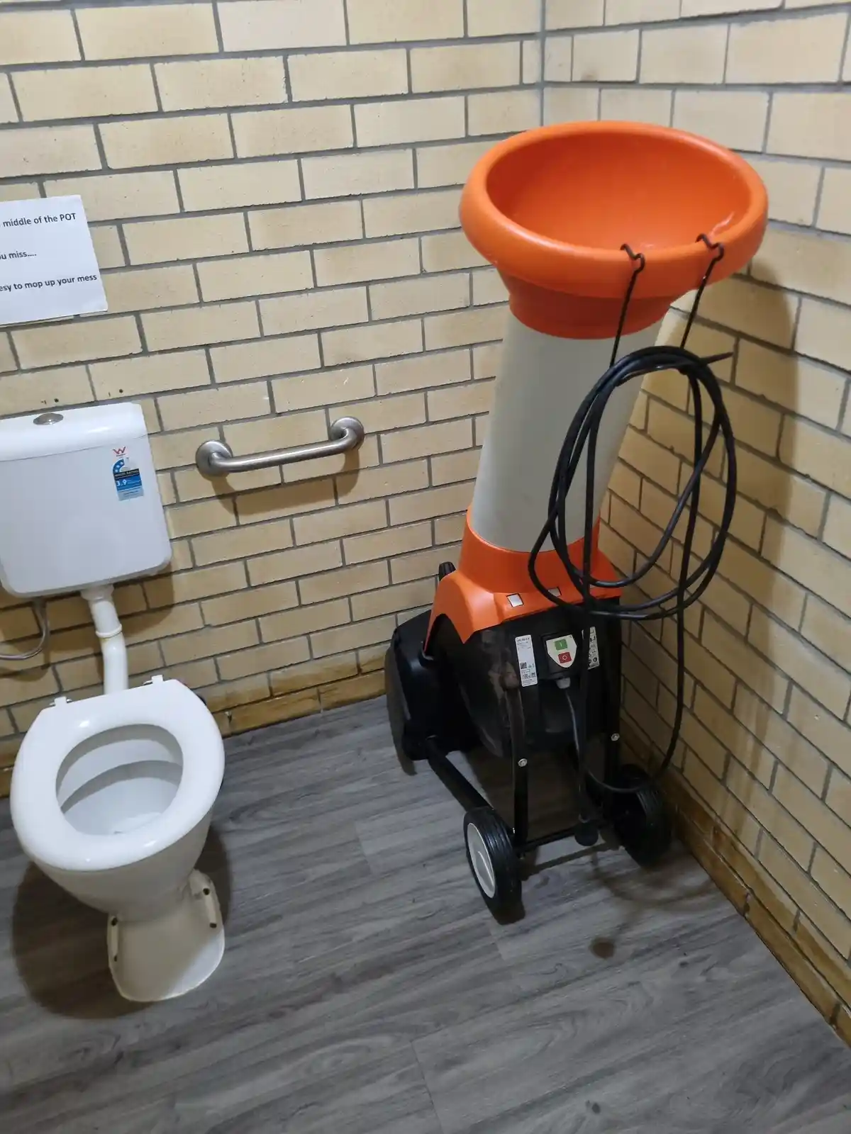 A bathroom stall with a white toilet and a beige brick wall, next to a large orange and white industrial floor cleaning…