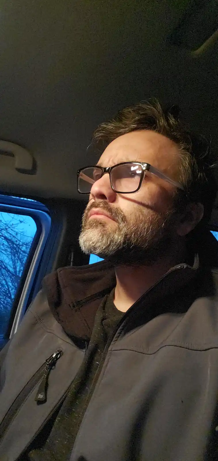 A man with glasses and a beard sits in a car at night, looking upward with a weary, unenthusiastic expression. The overlaid…