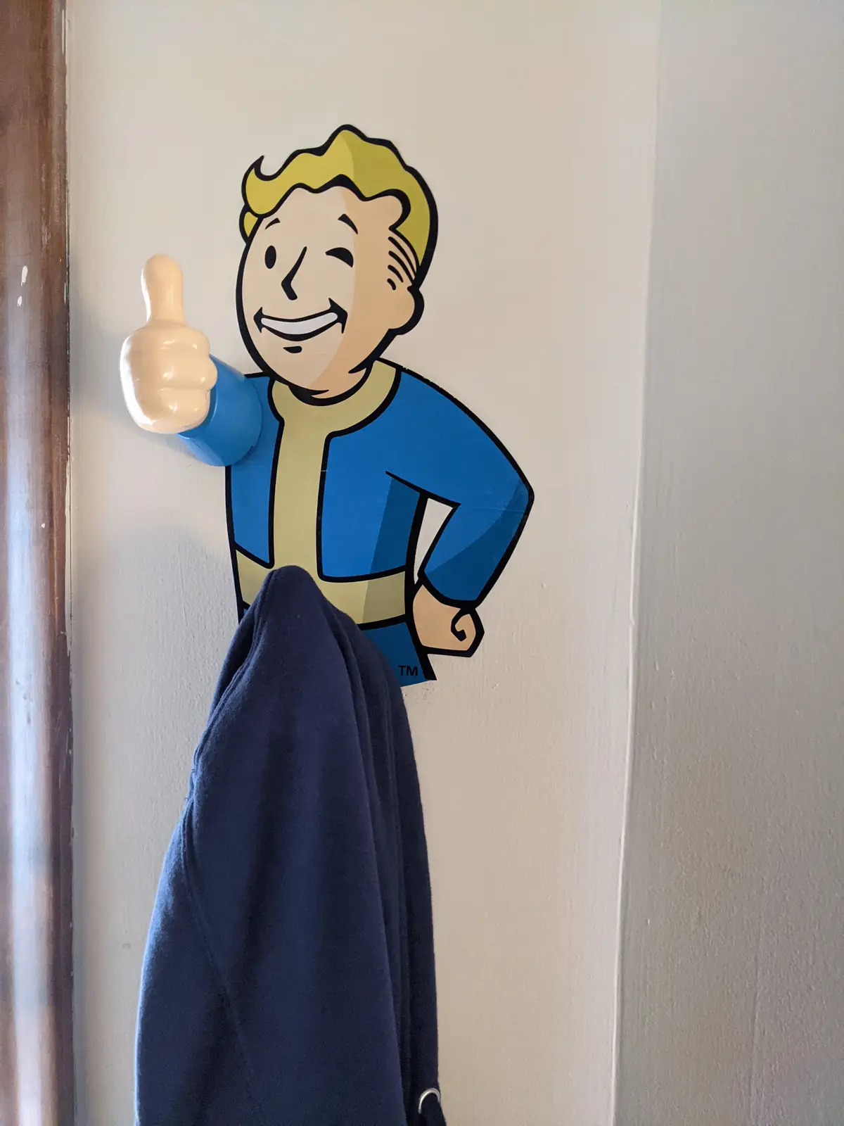 A blue jacket hangs from the thumb of a three-dimensional Fallout Vault Boy wall decoration, which is mounted on a white…