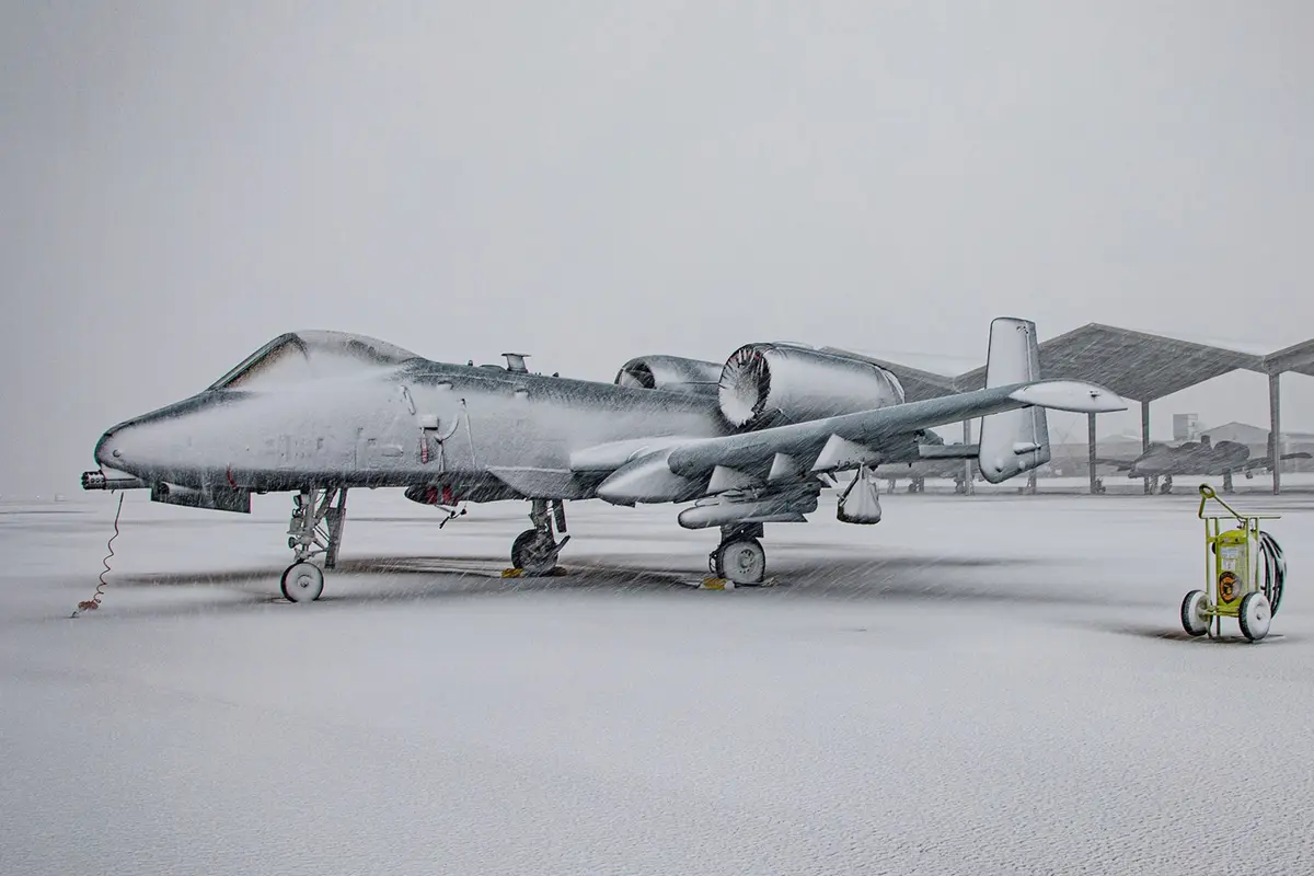A military fighter jet, covered in snow, sits on an airfield during a heavy snowstorm. The aircraft's fuselage, wings, and…