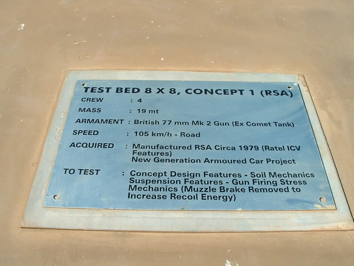 A blue metal plaque mounted on a sandy surface details a military vehicle called 'Test Bed 8x8, Concept 1 (RSA)'. It lists…