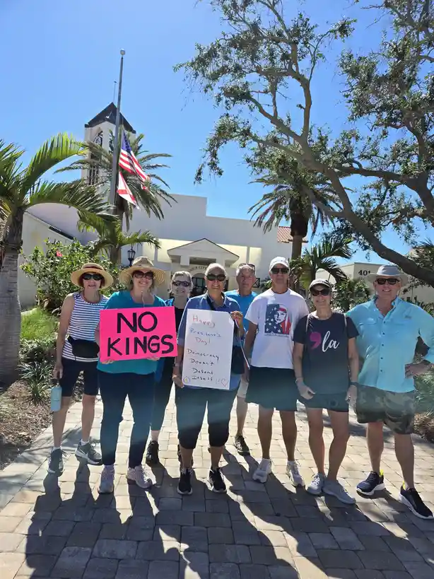 A group of smiling people stand outdoors in front of a building with a flagpole and palm trees, holding signs. One pink sign…