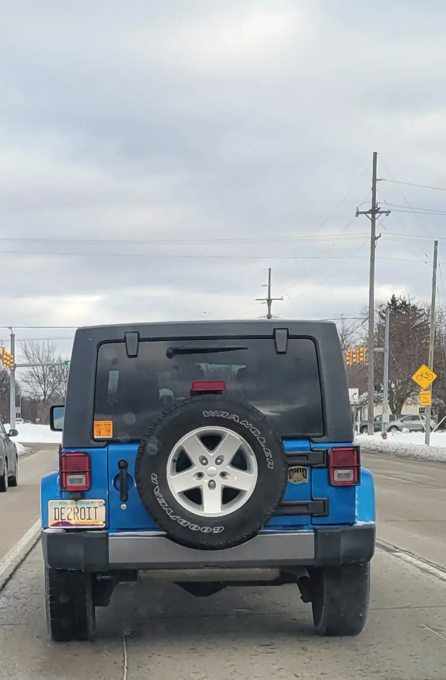 The rear view of a blue Jeep Wrangler with a spare tire mounted on the back, showing a Michigan license plate that reads…