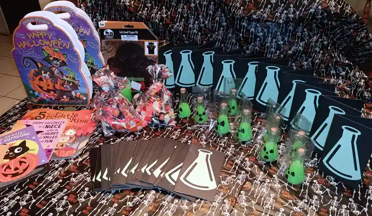 A table is covered in Halloween-themed party supplies, including skeleton-patterned tablecloths, Halloween coloring books…
