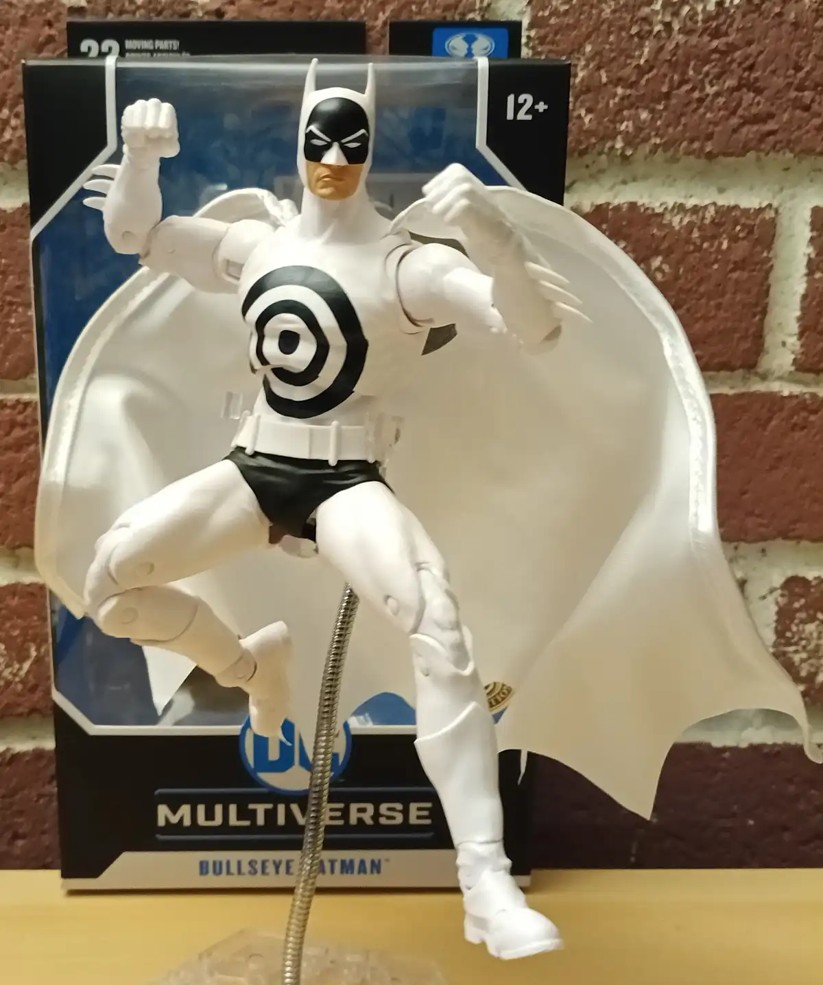 A white action figure of Batman, labeled 'Bullseye Batman' on its DC Multiverse packaging, stands in a dynamic pose with a…