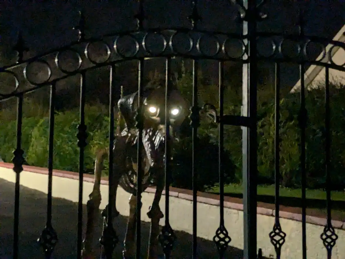 A nighttime photo shows a dark, ornate metal fence with a skeletal figure behind it, its glowing eyes reflecting light. The…