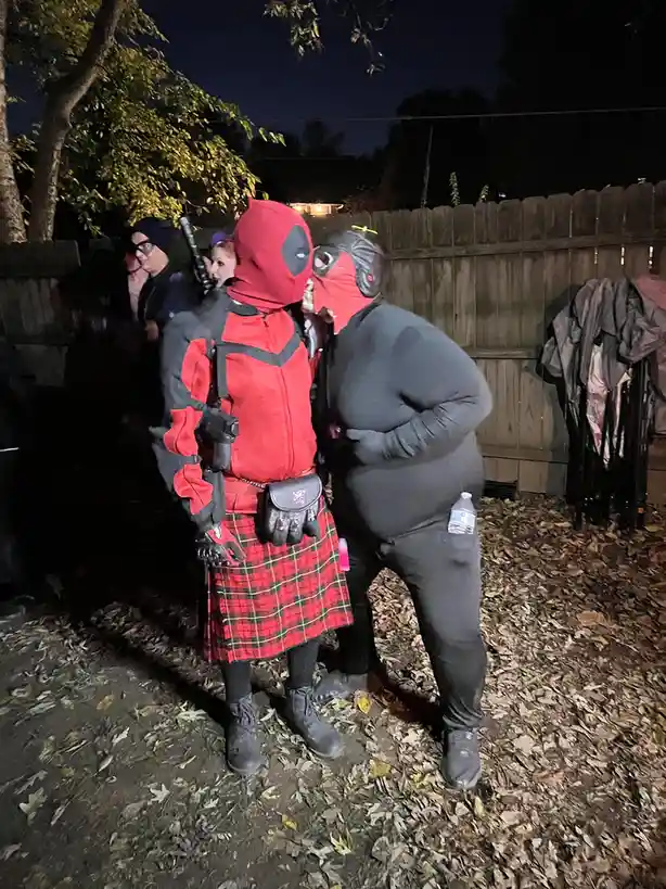 Two people in Deadpool costumes stand close together at night outdoors, one wearing a red and black Deadpool bodysuit with a…