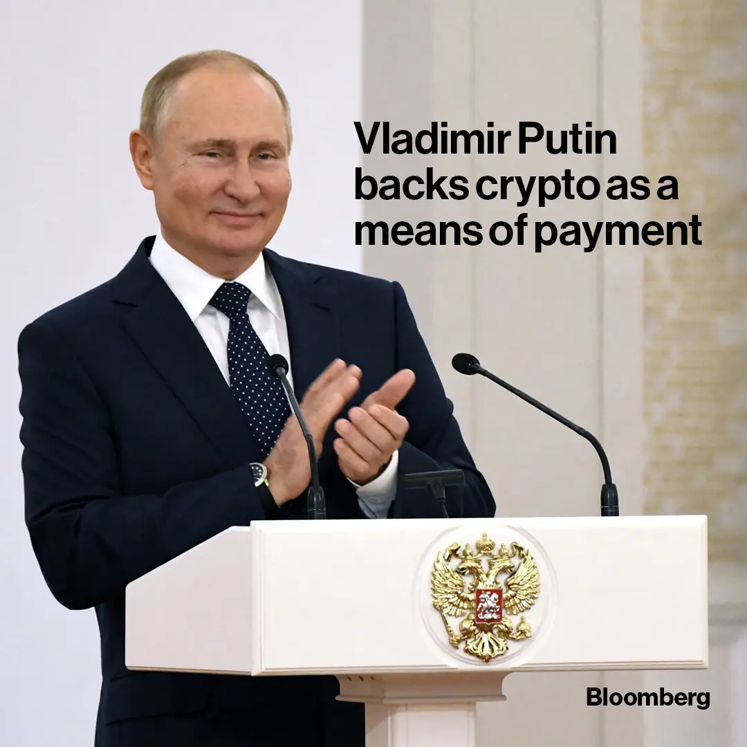 A man in a dark suit and tie stands behind a white podium with a golden Russian coat of arms, clapping his hands. Two…