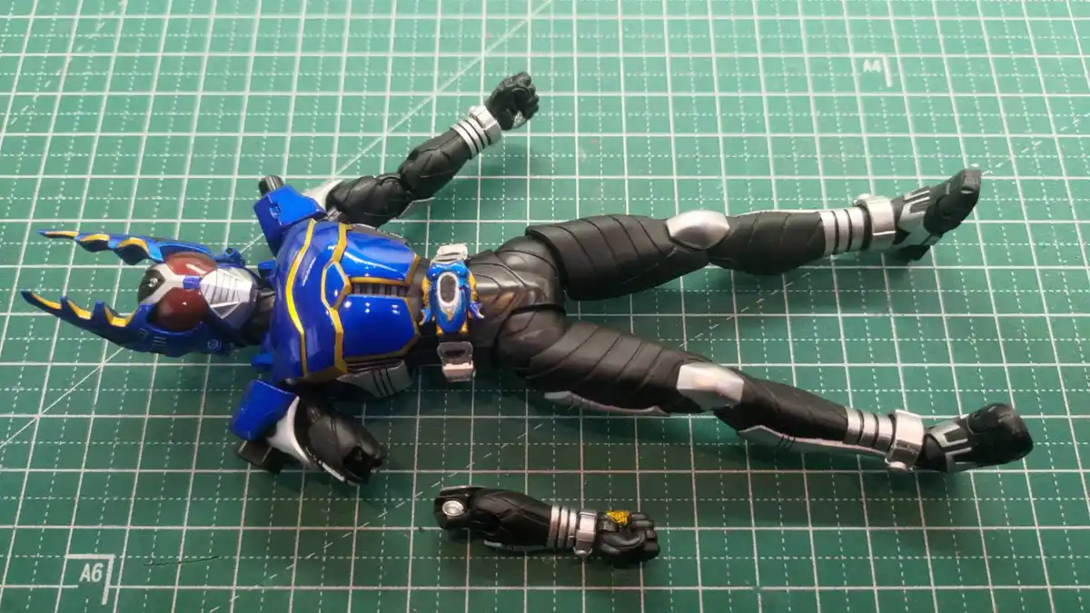A blue and black armored action figure, resembling a kamen rider, lies flat on its back on a green grid cutting mat with one…