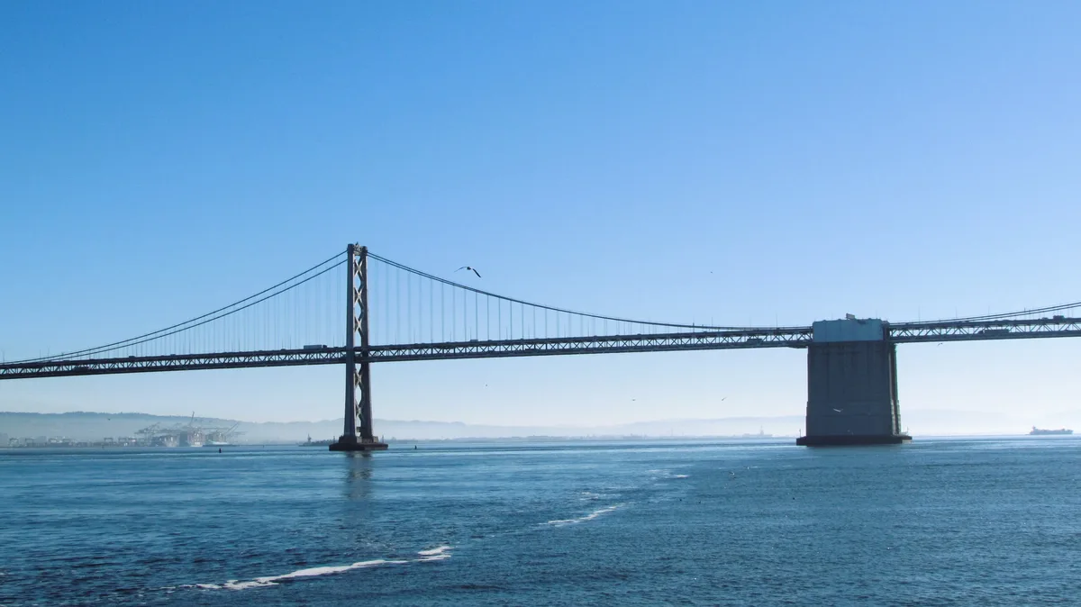 A wide-angle photograph shows the Bay Bridge spanning a large body of water under a clear blue sky. The bridge's steel…