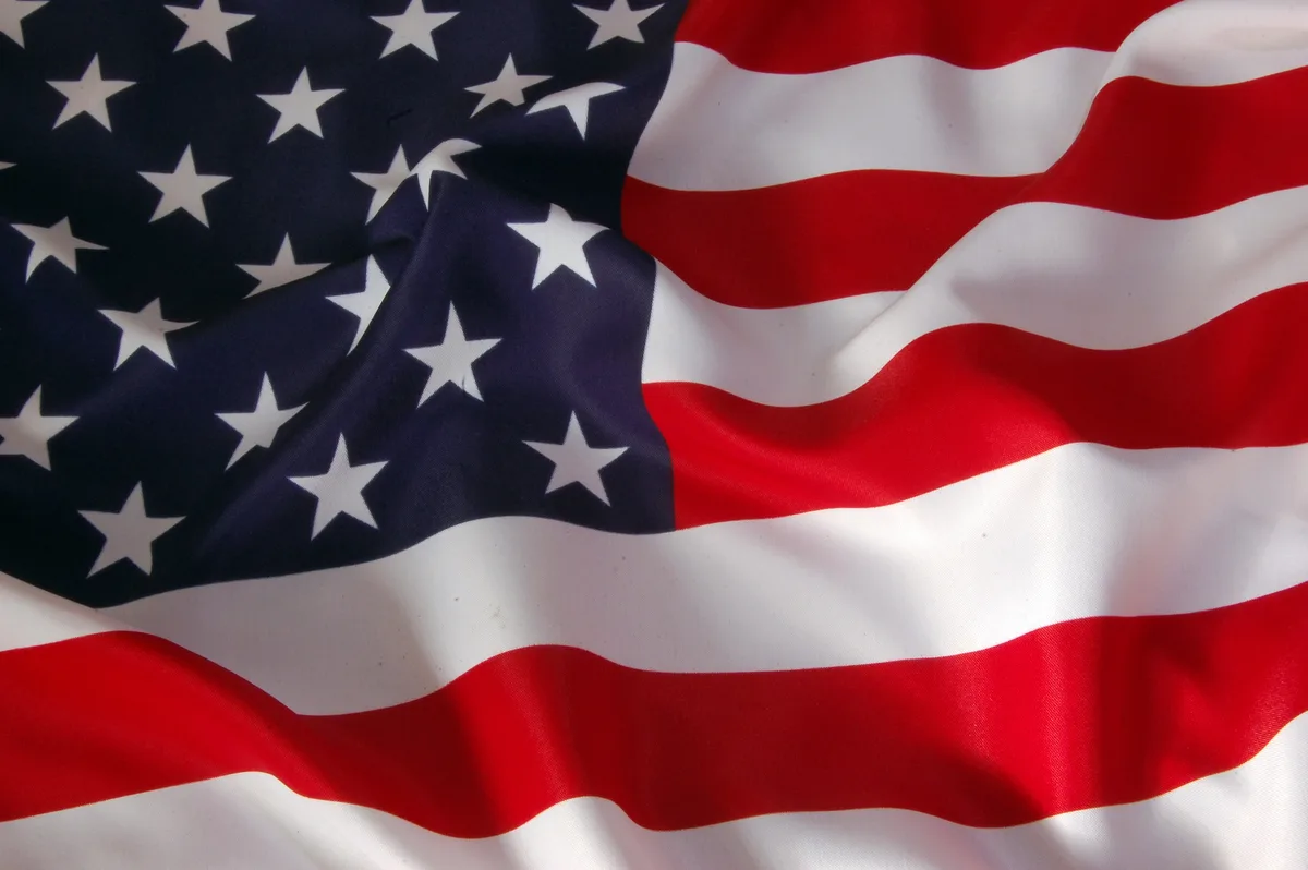 A close-up photograph of a waving American flag, showing its red and white stripes and blue canton with white stars. The…