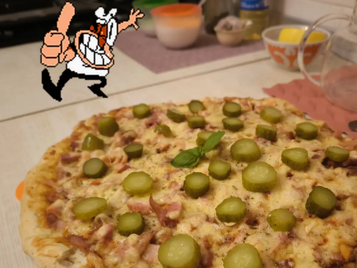 A homemade pizza covered in melted cheese, ham, and numerous pickle slices sits on a kitchen counter, with a pixelated chef…