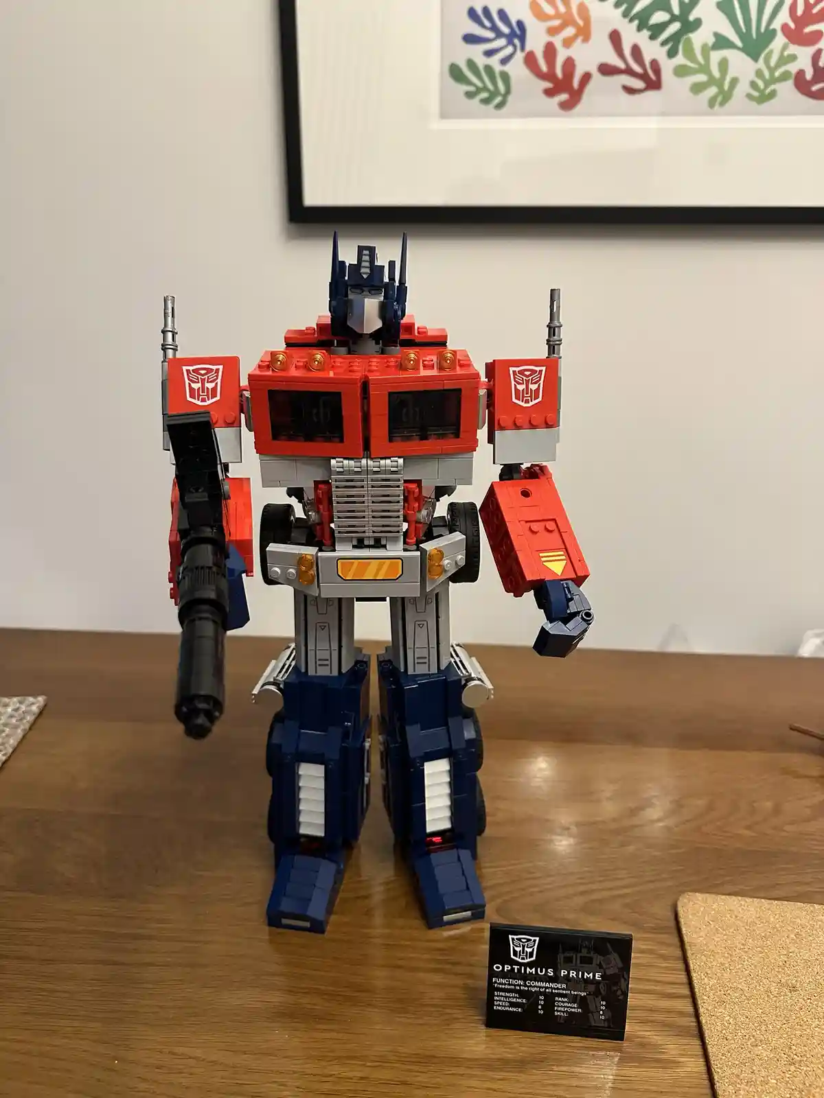 A large, red and blue LEGO Optimus Prime figure stands on a wooden table, holding a black cannon in one hand. A small black…